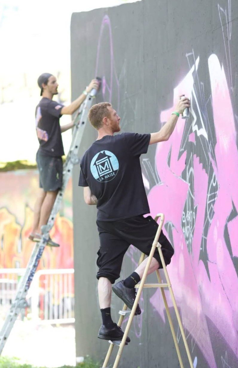 Deux artistes de La Maise, sur des échelles, peignent un graff rose sur un grand mur