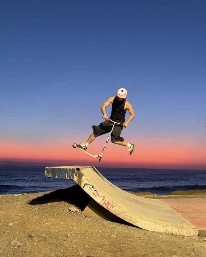 Timothé Faquin, casque rose, en plein saut en trottinette au-dessus d'une rampe, face à la mer au coucher du soleil